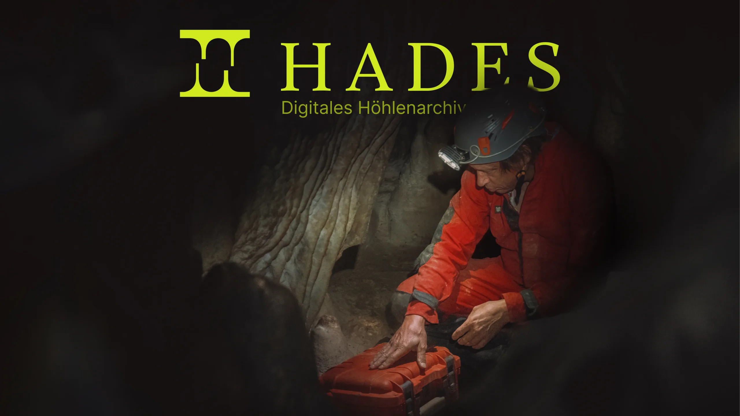 A caver in an orange suit and helmet lamp kneeling in a cave, under the title "HADES — Digitales Höhlenarchiv"
