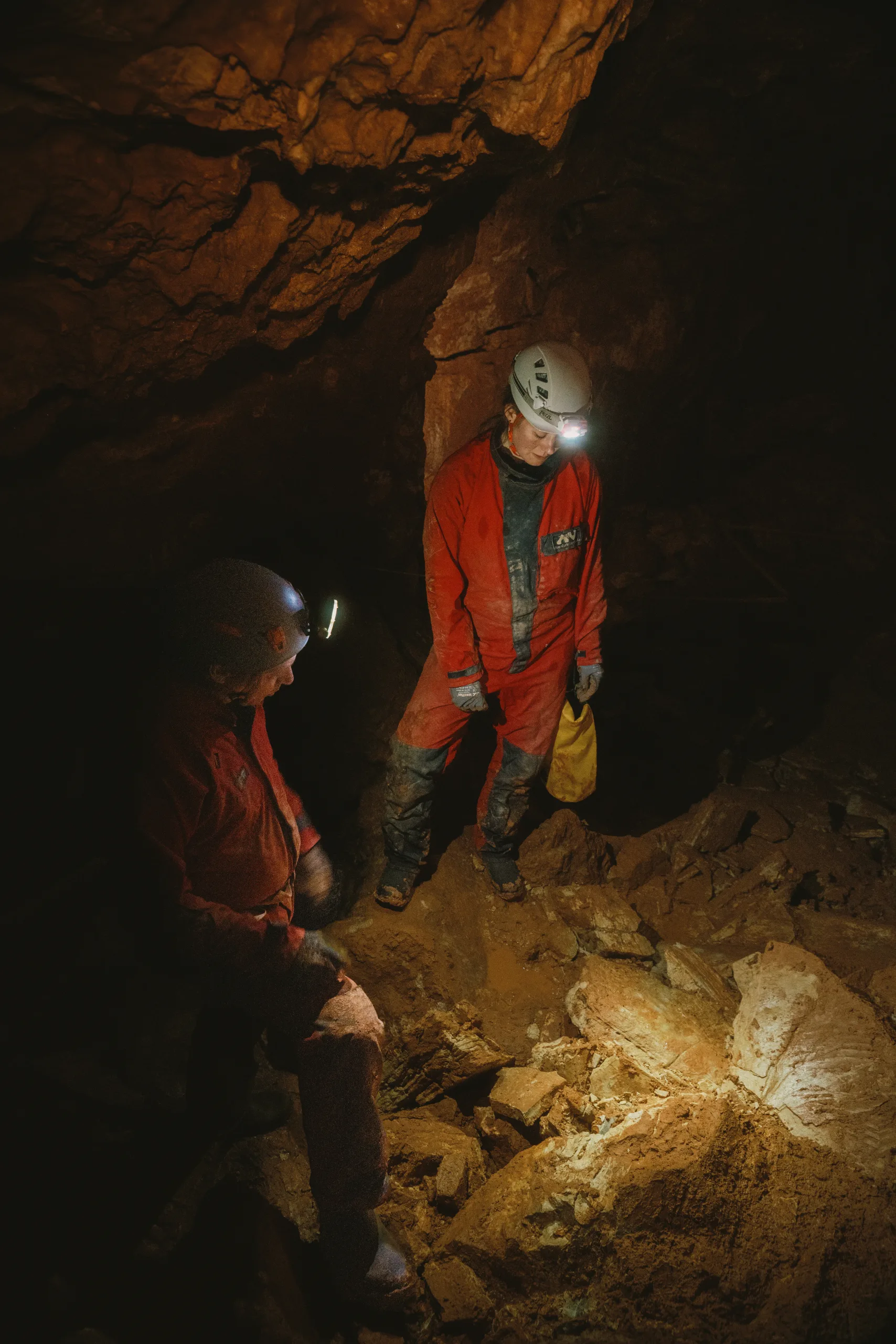 Two cavers with helmet lamps in a cave, one crouching by the rock, one standing behind