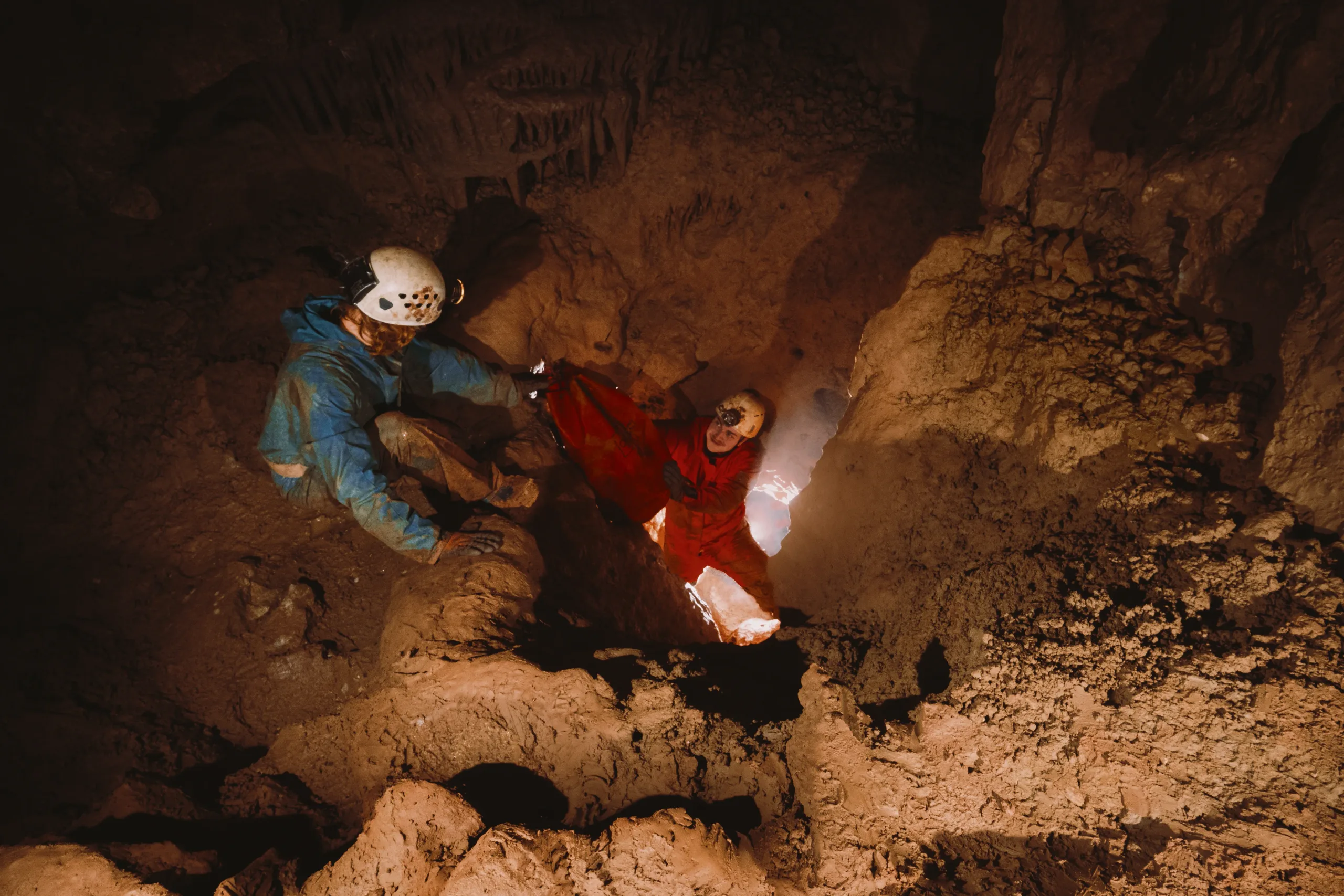 Two cavers in blue and red suits crouching between rocks, lit by a helmet lamp